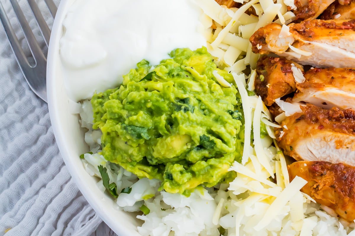 A close-up of a bowl with sliced chicken, shredded cheese, keto guacamole, white rice, and a dollop of sour cream.