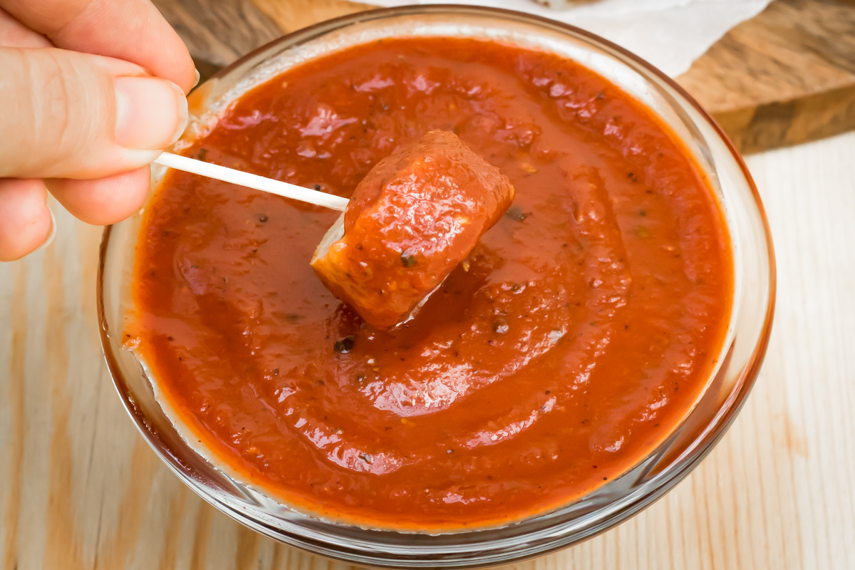 A hand holds a toothpick with a cube of food dipped in a bowl of red tomato sauce which is sugar free keto ketchup.