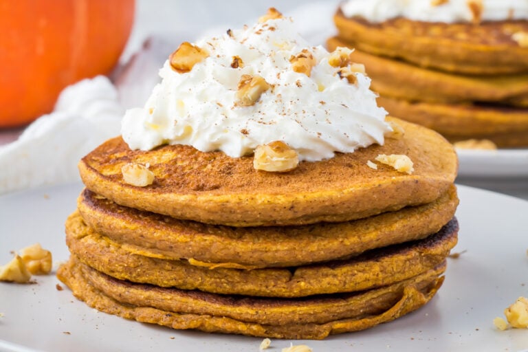A stack of whole30 pumpkin pancakes topped with whipped coconut cream and chopped walnuts on a white plate.