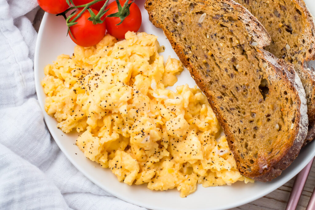 Plate with scrambled eggs topped with black pepper, two slices of multigrain toast, and a cluster of vine tomatoes.