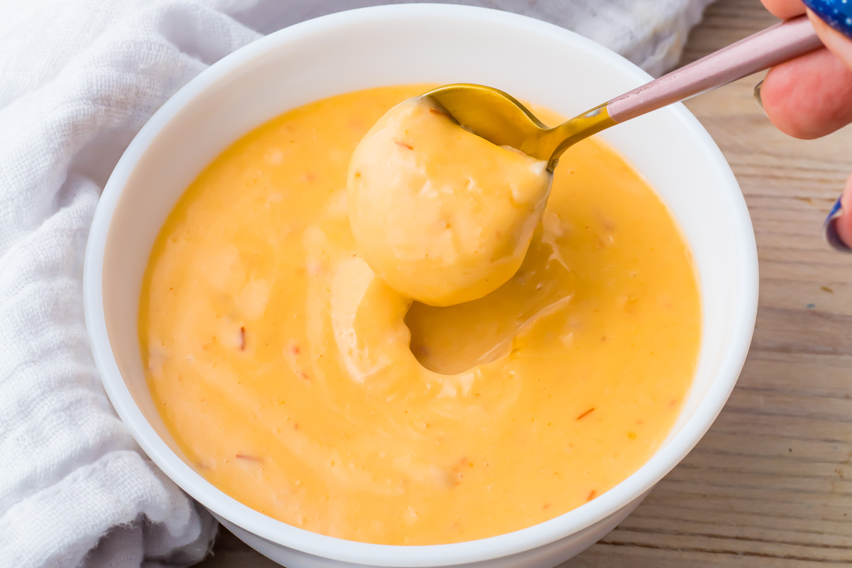A spoon scooping creamy, yellow spicy sriracha without mayo from a white bowl on a wooden surface next to a white cloth&mdash;perfect for your Game Day Recipes lineup.