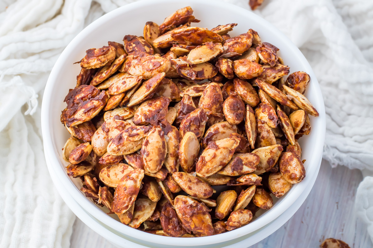 A white bowl filled with roasted sweet pumpkin seeds coated in seasoning,  placed on a light-colored surface with a textured white fabric nearby.