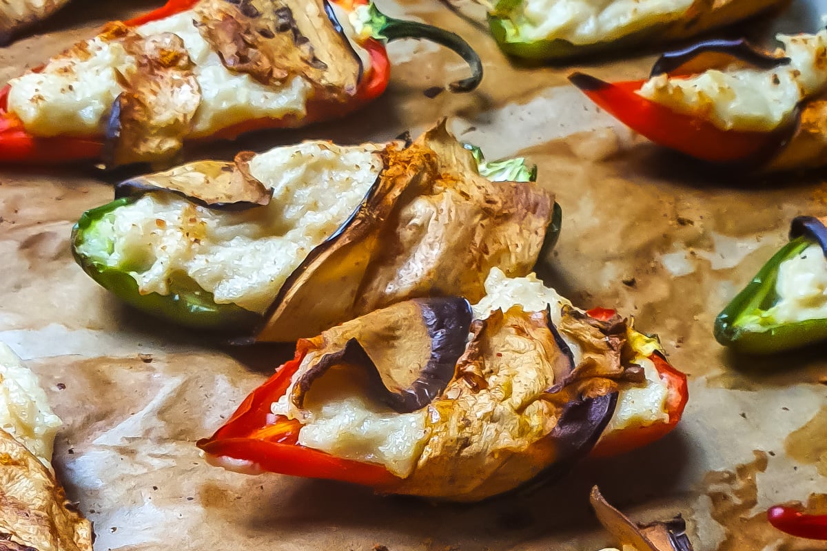 Stuffed red and green jalapeno peppers, wrapped with thin slices of roasted eggplant, arranged on a parchment-lined baking sheet for vegan jalapeno poppers.