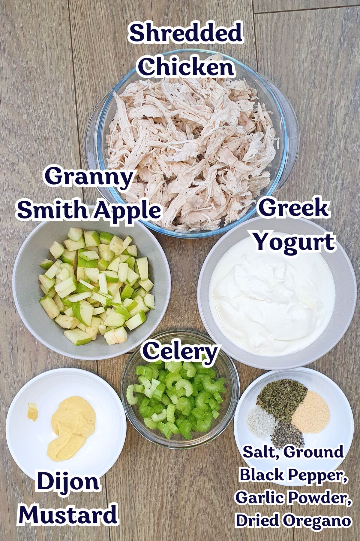 Bowls containing shredded chicken, chopped Granny Smith apple, sliced celery, Greek yogurt, Dijon mustard, and a mix of salt, black pepper, garlic powder, and dried oregano on a wooden surface.