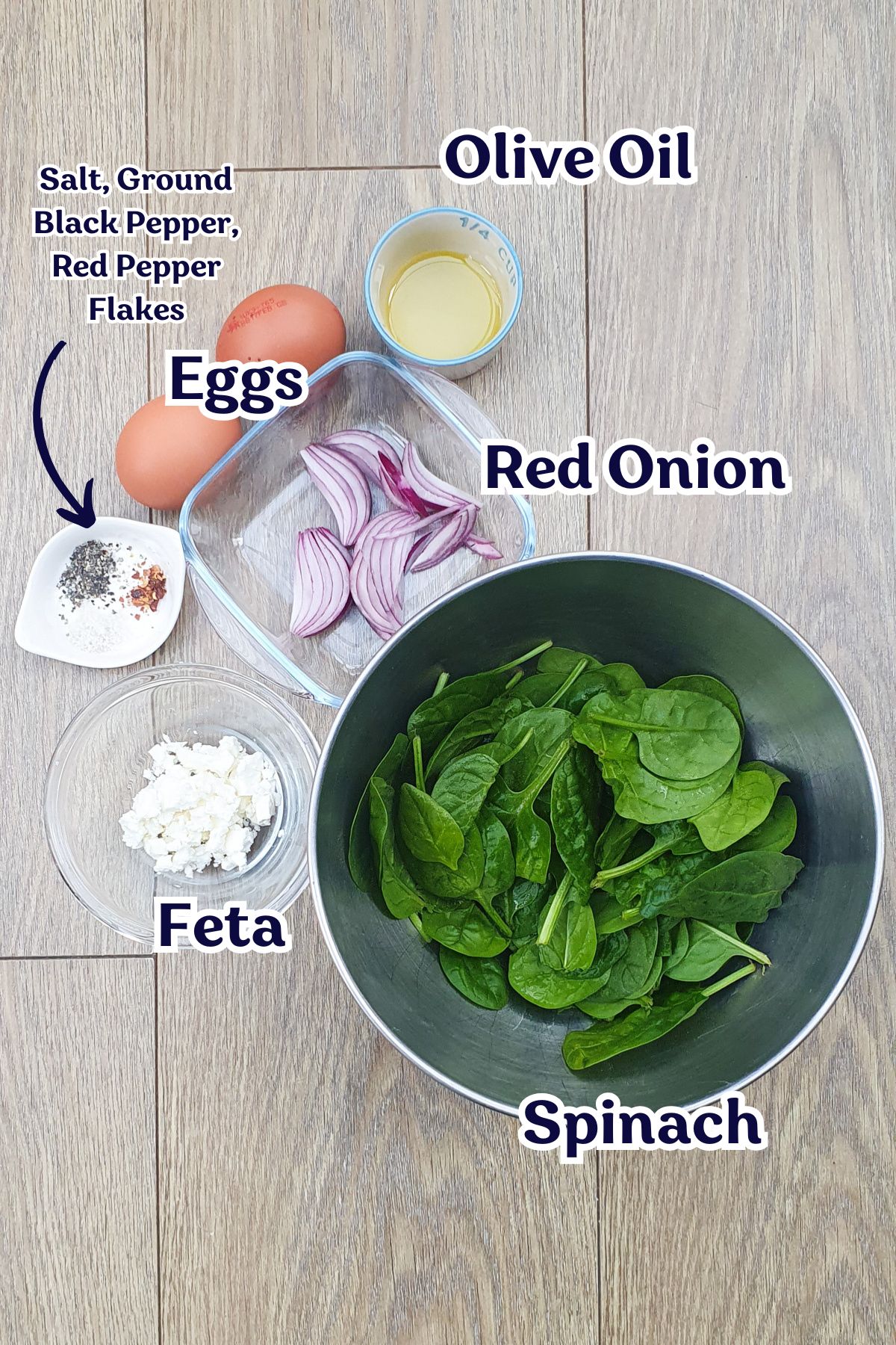 A display of ingredients on a wooden surface: fresh spinach in a bowl, sliced red onion, eggs, olive oil, feta cheese, and a small dish of salt, black pepper, and red pepper flakes.