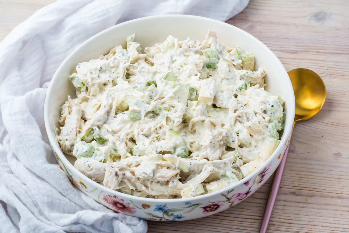 A bowl of Greek yogurt chicken salad with chopped celery and creamy dressing sits on a wooden table next to a gold spoon and a white cloth.