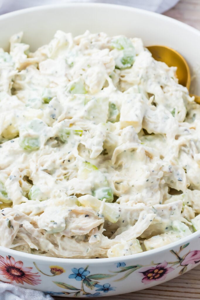 A close-up of a bowl filled with creamy Greek yogurt chicken salad mixed with celery and herbs, with a serving spoon partially visible.