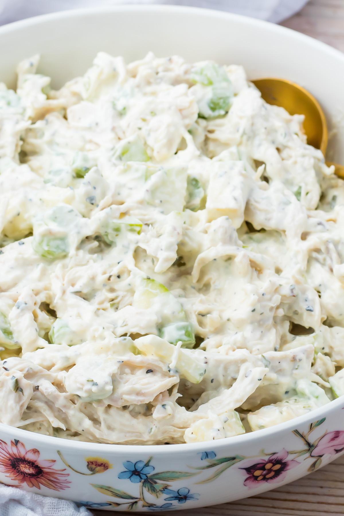 A close-up of a bowl filled with creamy Greek yogurt chicken salad mixed with celery and herbs, with a serving spoon partially visible.