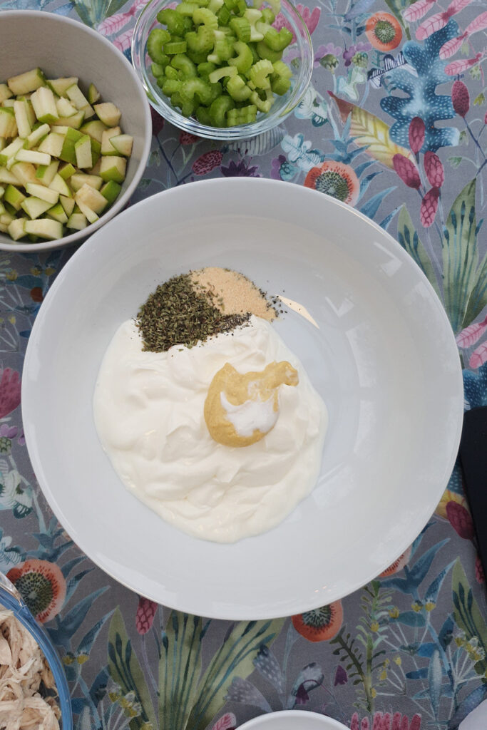 A white bowl with Greek yogurt, mustard, dried herbs, and garlic powder sits on a table next to bowls of chopped celery and green apples.