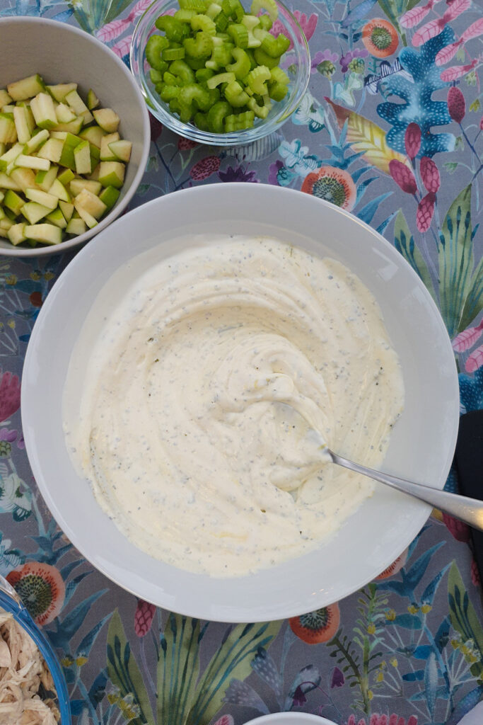 A bowl of creamy dressing with a spoon, surrounded by bowls of chopped green apple and sliced celery on a floral-patterned tablecloth.