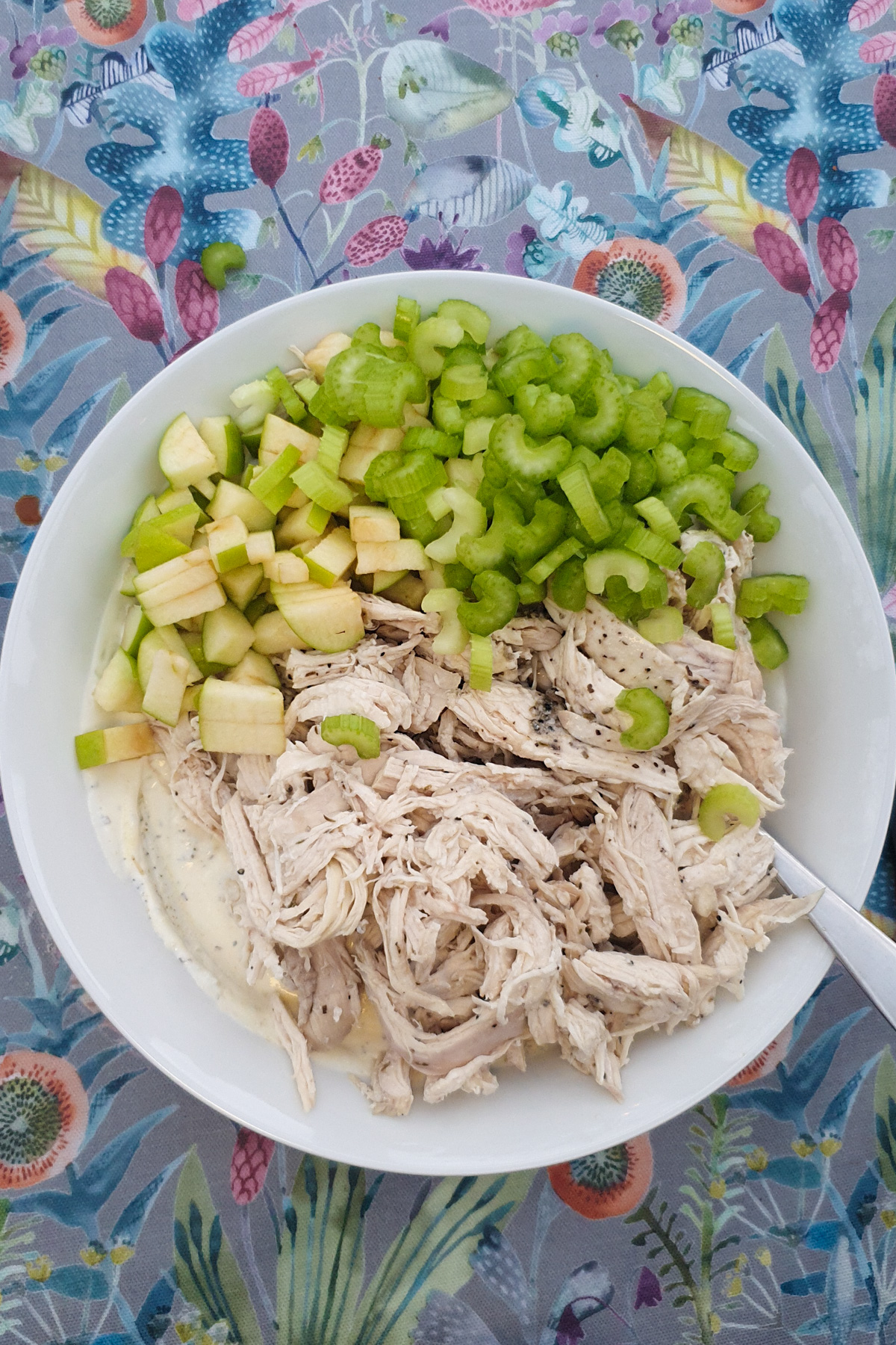 A white bowl with shredded chicken, diced green apple, chopped celery, and Greek yogurt dressing sits on a colorful floral tablecloth.