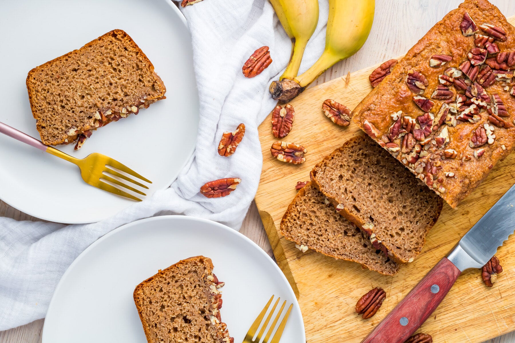 A loaf of healthy banana bread topped with pecans is sliced on a wooden board, with two plated slices and gold forks beside scattered pecans and bananas.