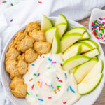 A plate with green apple slices, animal-shaped cookies, and a swirl of healthy dunkaroo dip topped with colorful sprinkles. A small bowl of sprinkles is nearby.