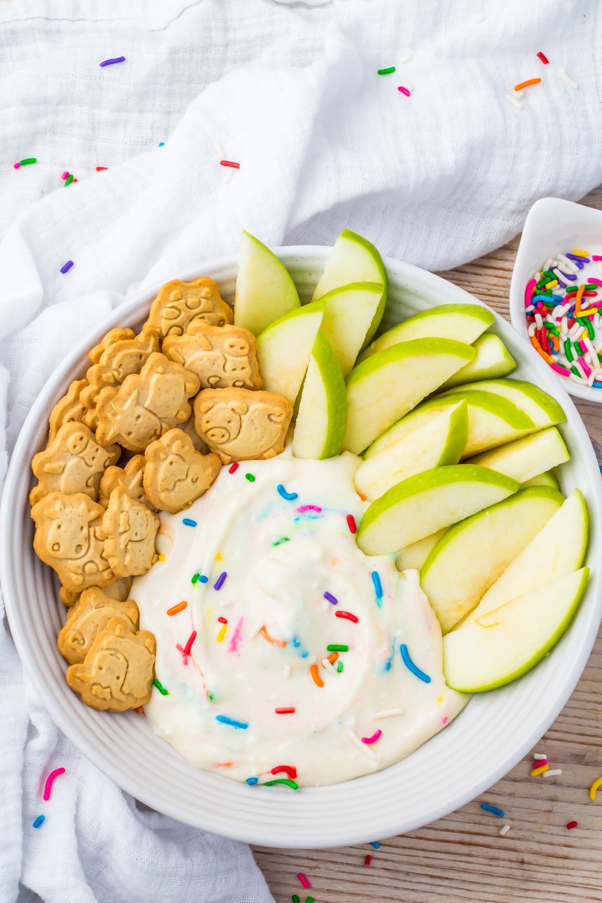 A plate with green apple slices, animal-shaped cookies, and a swirl of healthy dunkaroo dip topped with colorful sprinkles. A small bowl of sprinkles is nearby.