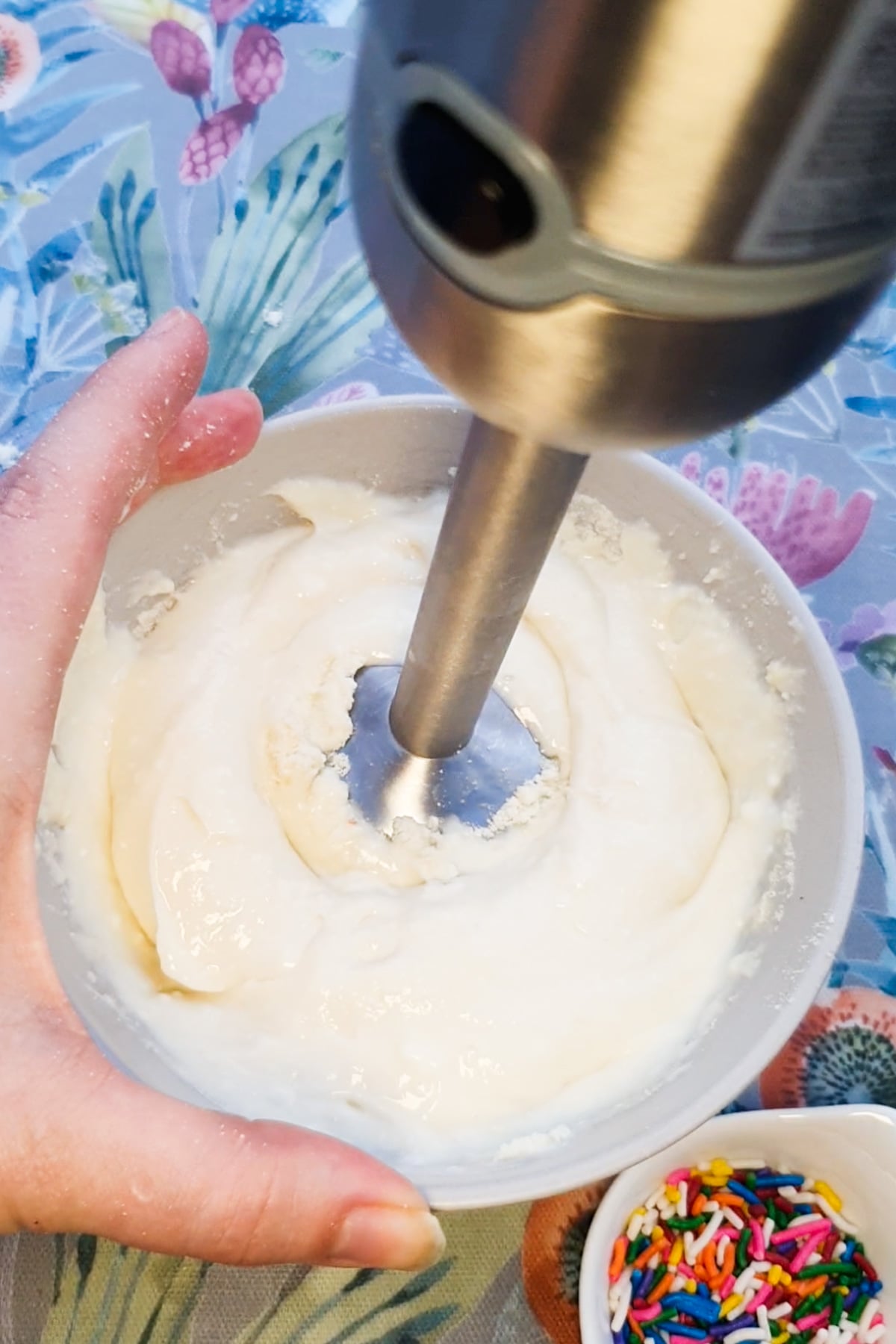 A hand holds a bowl of thick white mixture while blending with a hand blender; a small bowl of colorful sprinkles is nearby on a patterned tablecloth.