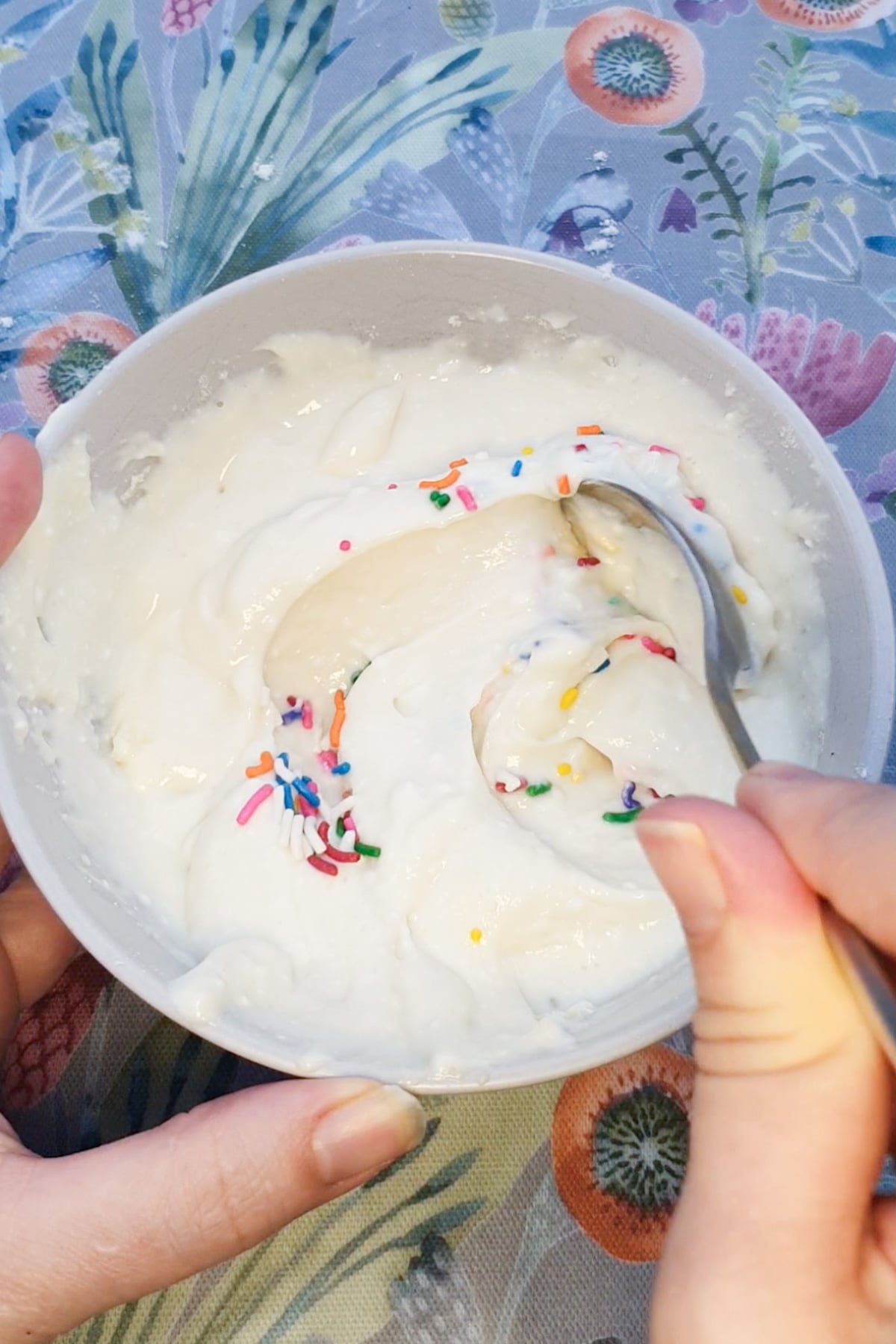 A person stirs white creamy dunkaroo dip with colorful sprinkles in a bowl, using a spoon, on a table with a floral patterned cloth.