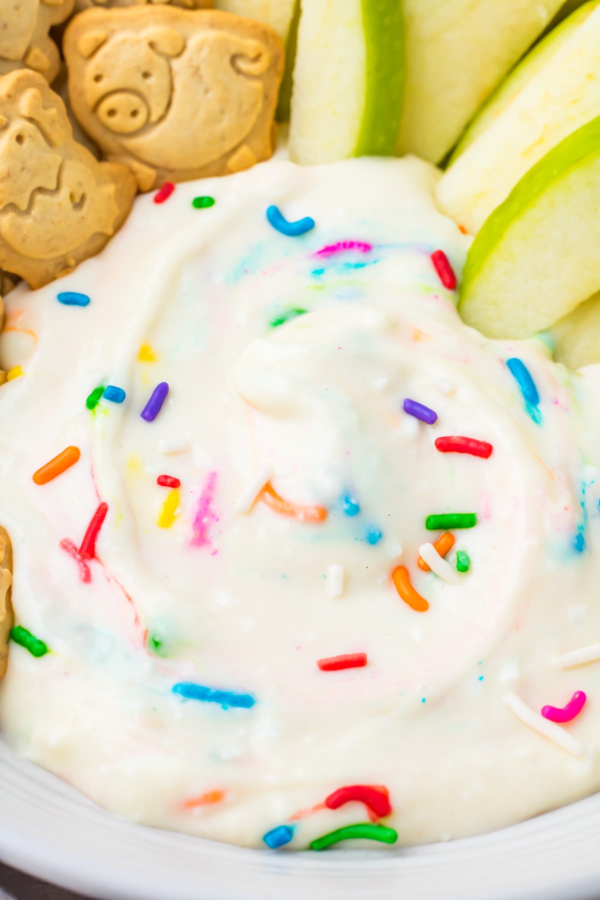 A close-up of healthy dunkaroo dip with colorful sprinkles, surrounded by green apple slices and animal-shaped cookies.