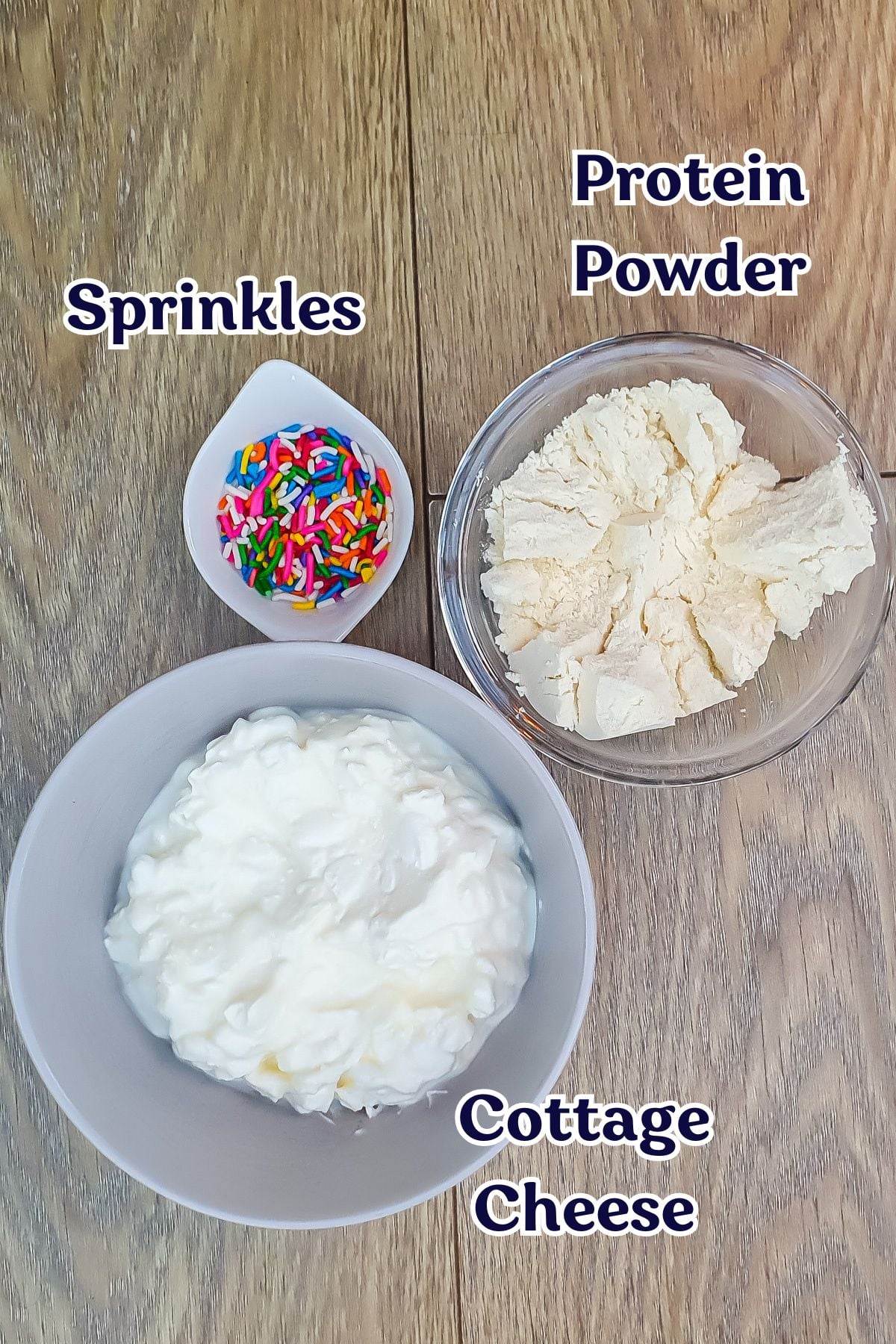 A bowl of cottage cheese, a bowl of protein powder, and a small dish of colorful sprinkles are arranged on a wooden surface, each labeled accordingly.