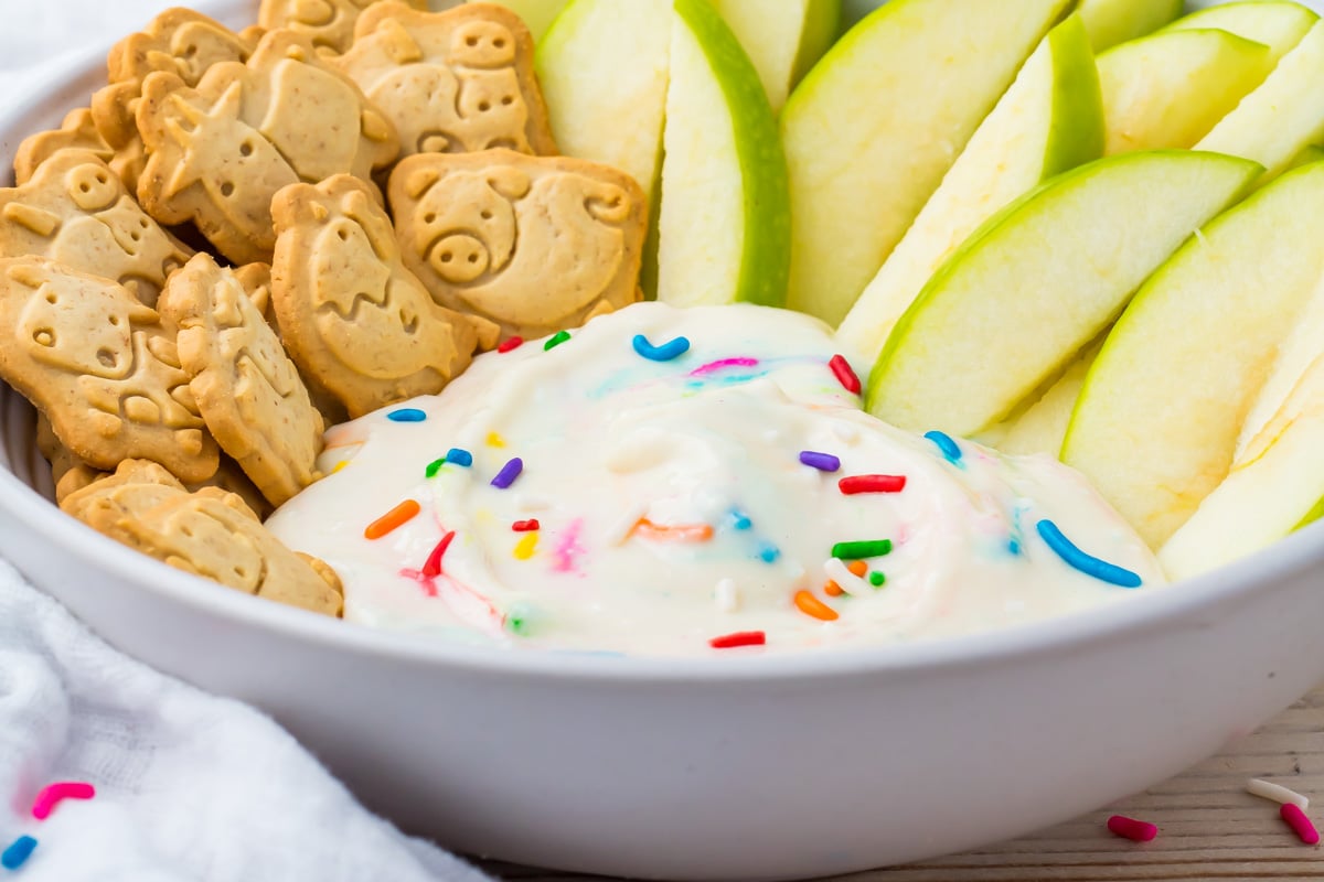 A bowl containing animal-shaped cookies, green apple slices, and a swirl of healthy dunkaroo dip topped with colorful sprinkles.