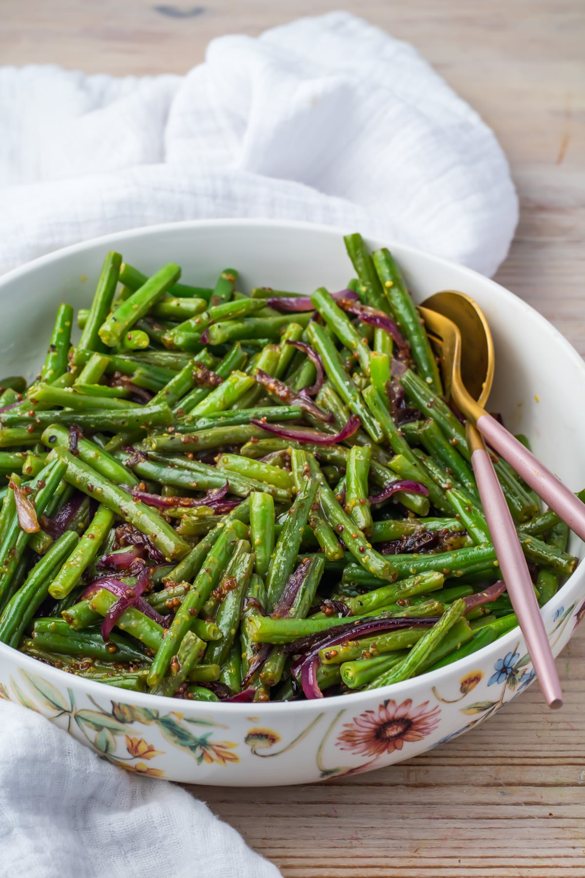 A bowl of honey mustard green beans mixed with sliced red onions, served with a gold spoon and pink fork on a light wooden table.