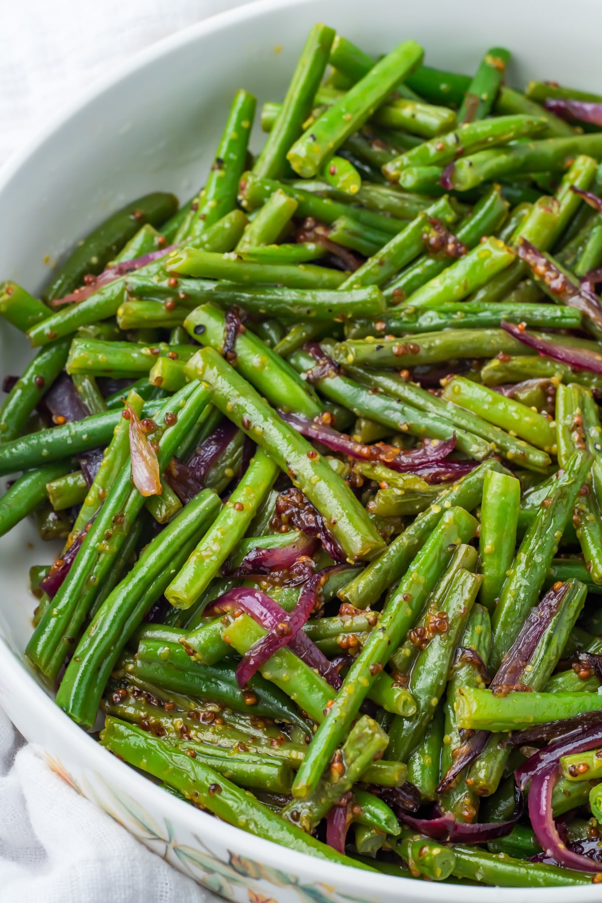 A bowl of cooked honey mustard green beans mixed with sliced red onions and a grainy mustard dressing.