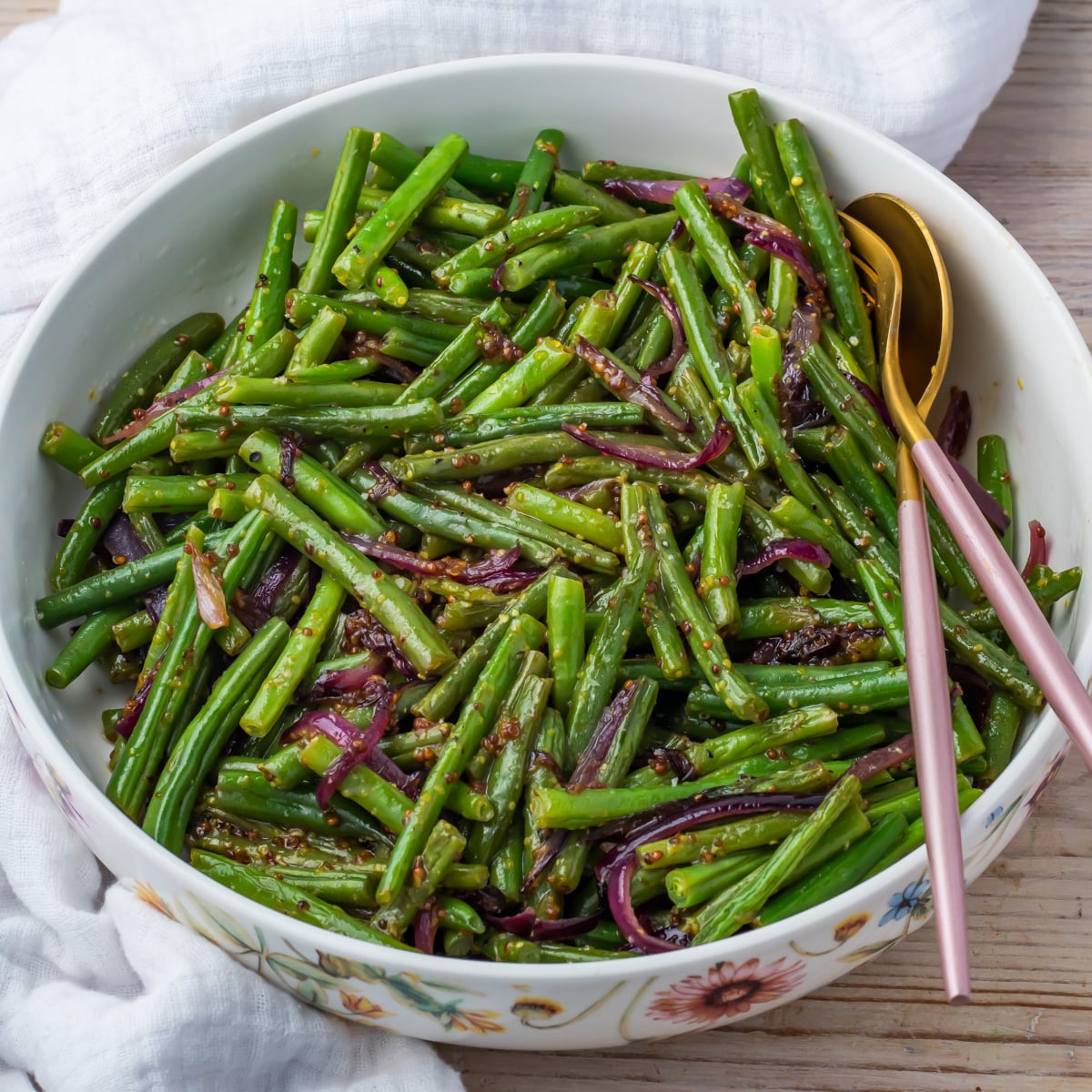 A bowl of sautéed honey mustard green beans and red onions mixed with seasoning, served with a gold fork and spoon on the side.