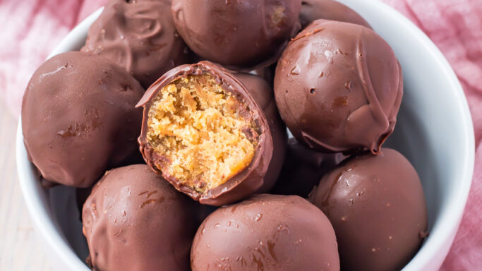 A white bowl filled with chocolate-covered peanut butter balls, one bitten to reveal the filling—an indulgent treat that could inspire healthy breakfast recipes. Pink cloth decorates the background.