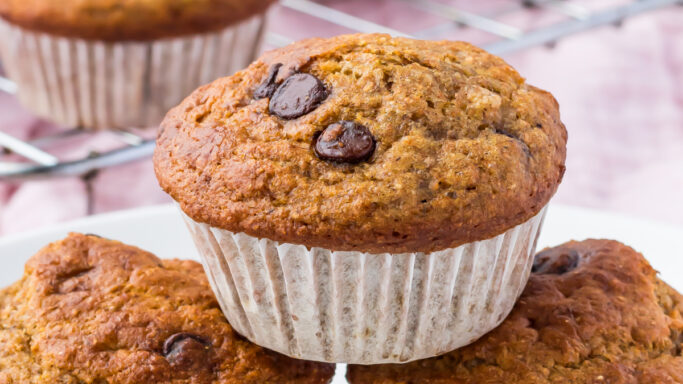 Three banana chocolate chip muffins on a white plate, with more muffins cooling on a wire rack in the background—perfect for those seeking healthy breakfast recipes.