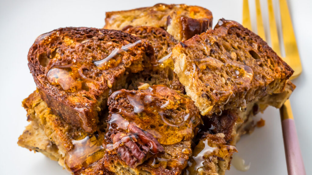 A serving of baked French toast with pecans and a drizzle of syrup sits on a white plate next to a gold fork, making it a delicious option for those seeking healthy breakfast recipes.