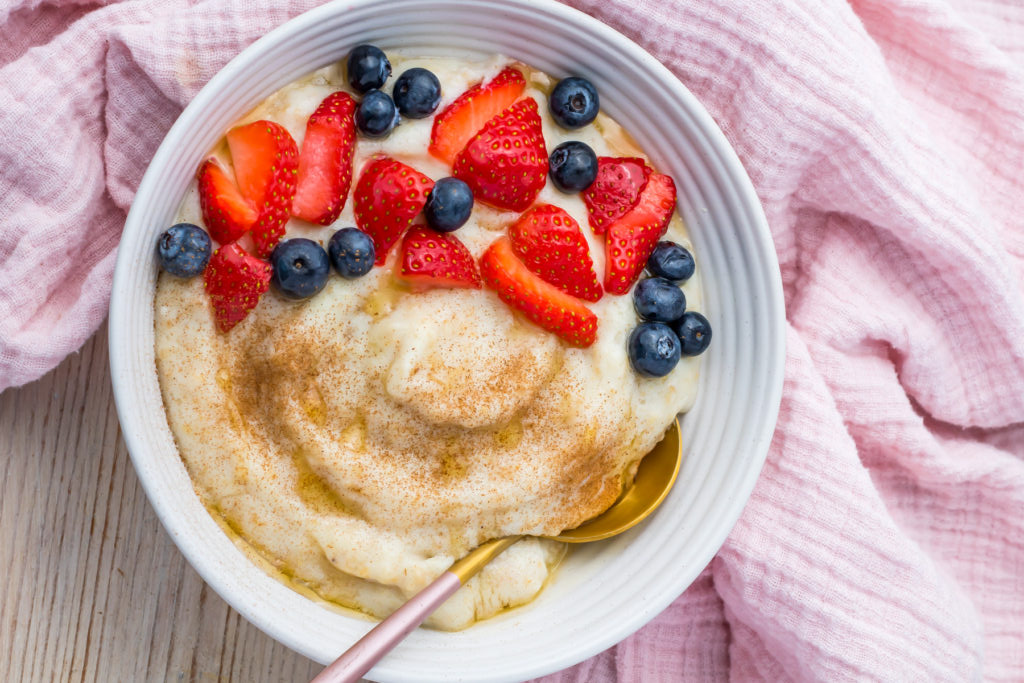 A white bowl of creamy porridge topped with sliced strawberries, blueberries, and a sprinkle of cinnamon sits with a gold spoon inside—perfect inspiration for healthy breakfast recipes. Pink fabric is in the background.