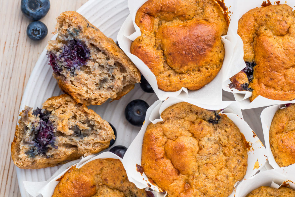 A close-up of blueberry muffins in white paper liners on a plate, with one muffin cut in half to reveal the blueberry filling. Fresh blueberries are scattered nearby—perfect inspiration for healthy breakfast recipes.