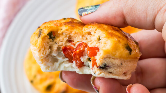 A hand holds a mini egg muffin with a bite taken out, showing a filling of red bell peppers and cheese—perfect for healthy breakfast recipes. More egg muffins rest on a white plate in the background.