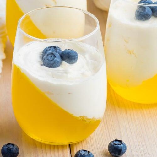A glass filled with yellow keto jello set at an angle, topped with a layer of white cream and garnished with blueberries; more identical keto desserts and scattered blueberries nearby.