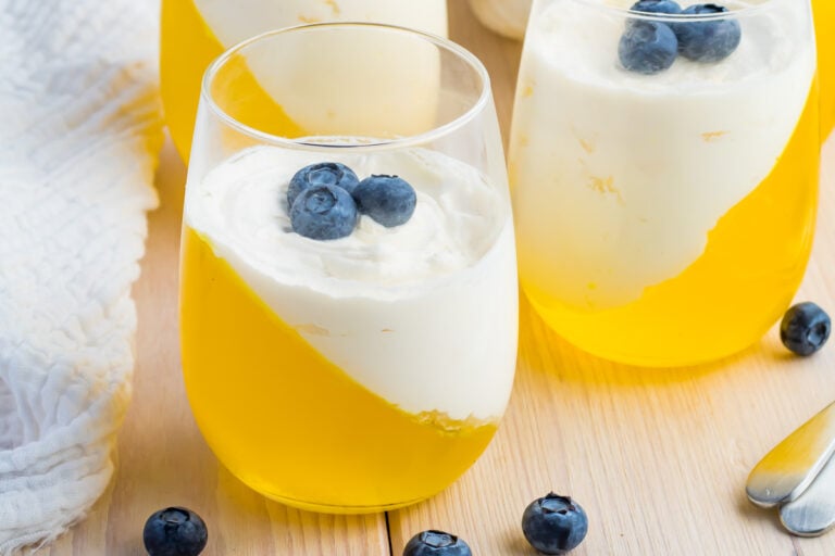 A glass filled with yellow keto jello set at an angle, topped with a layer of white cream and garnished with blueberries; more identical keto desserts and scattered blueberries nearby.