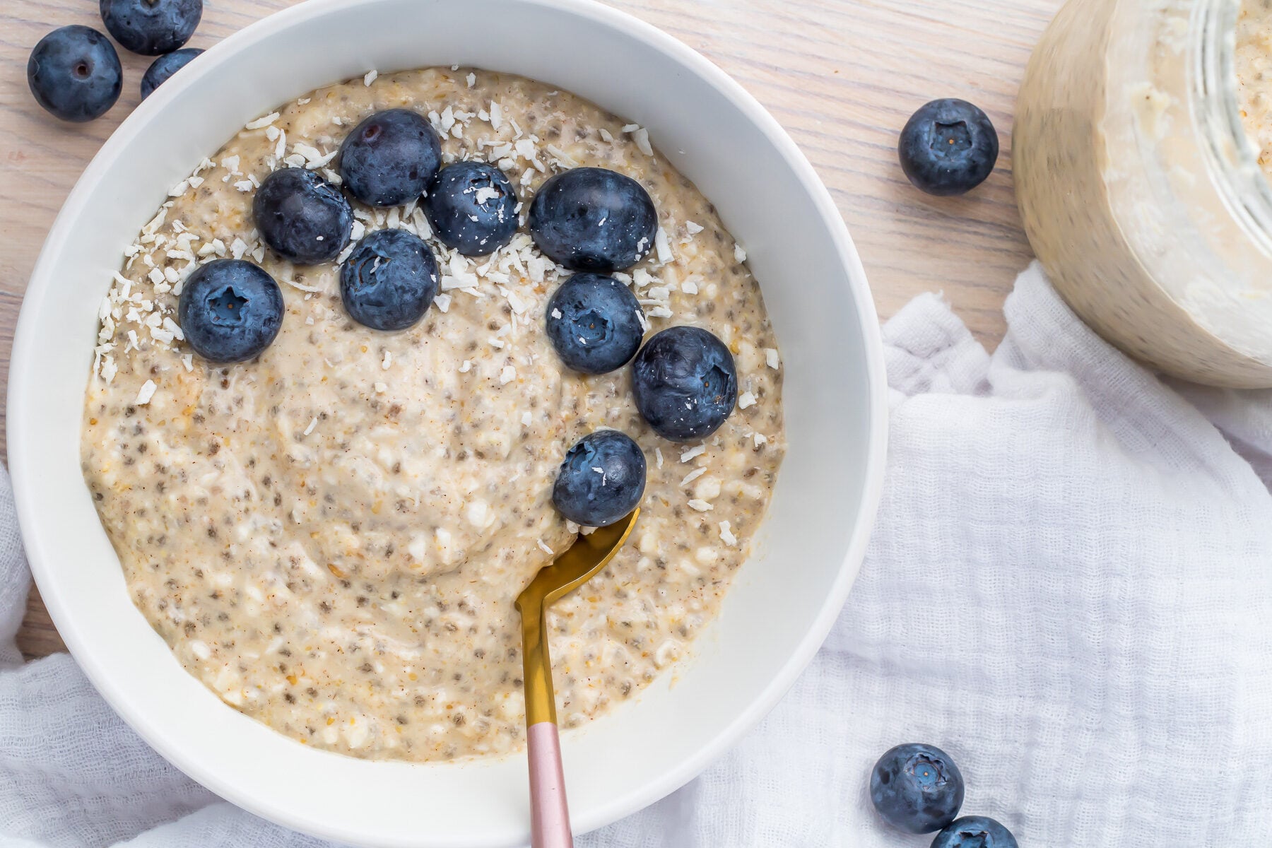 A bowl of keto overnight oats topped with fresh blueberries and shredded coconut, with a gold spoon inside. Additional blueberries are scattered on a wooden table beside a white cloth. A jar of oatmeal is partially visible in the background.