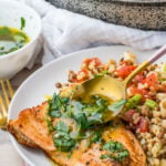 A plate of pan-seared salmon with skin topped with herb sauce, served with a side of grain salad. A spoon is pouring extra sauce over the salmon; a skillet with more fish is in the background.