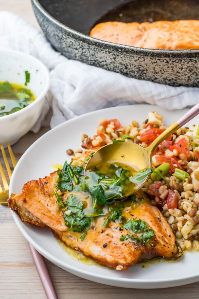 A plate of pan-seared salmon with skin topped with herb sauce, served with a side of grain salad. A spoon is pouring extra sauce over the salmon; a skillet with more fish is in the background.