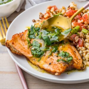 A plate with pan-seared salmon with skin topped with herb sauce, served with a side of mixed grains and diced vegetables, and a spoon drizzling sauce over the fish.