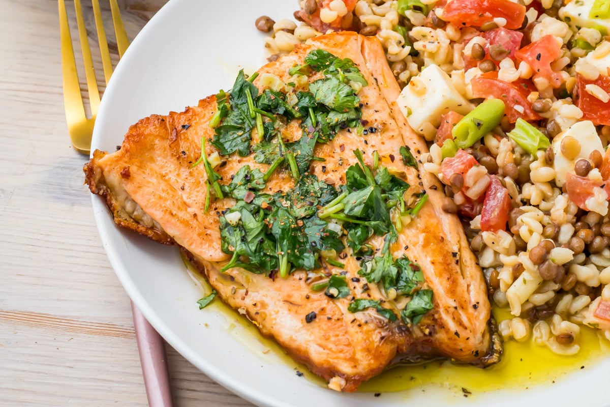 Pan-seared salmon fillet with skin topped with chopped herbs, served with a side of barley and lentil salad with tomatoes, green onions, and cheese on a white plate.