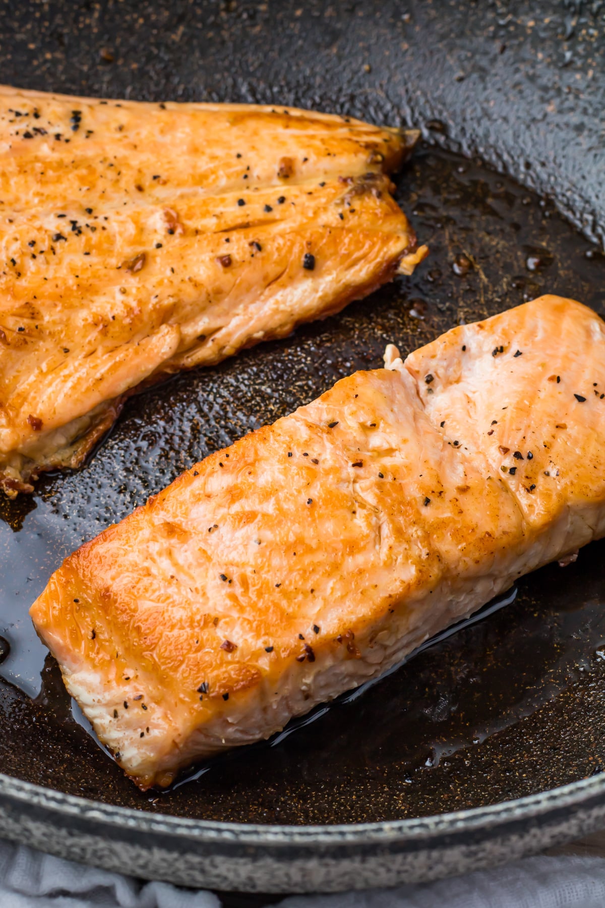 Two pieces of pan-seared salmon fillets with skin cooking in a black skillet, seasoned with salt and pepper.