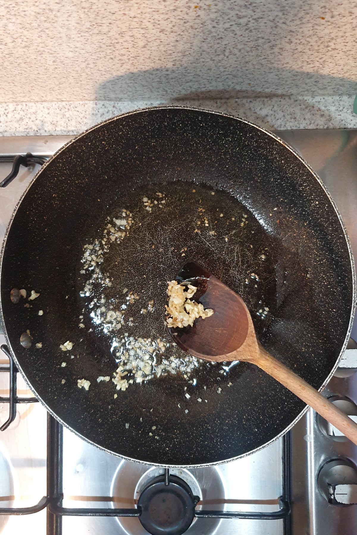 Minced garlic being saut&eacute;ed in a black frying pan with oil on a gas stove, stirred with a wooden spoon.