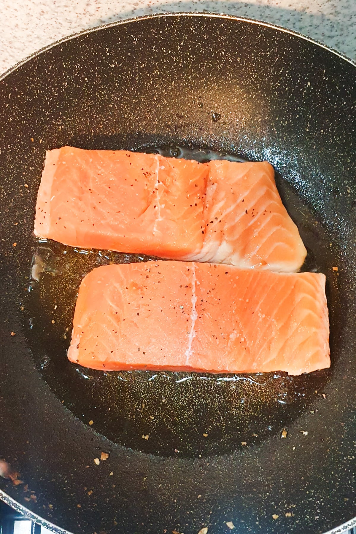 Two raw salmon fillets seasoned with pepper are cooking in oil in a nonstick frying pan.