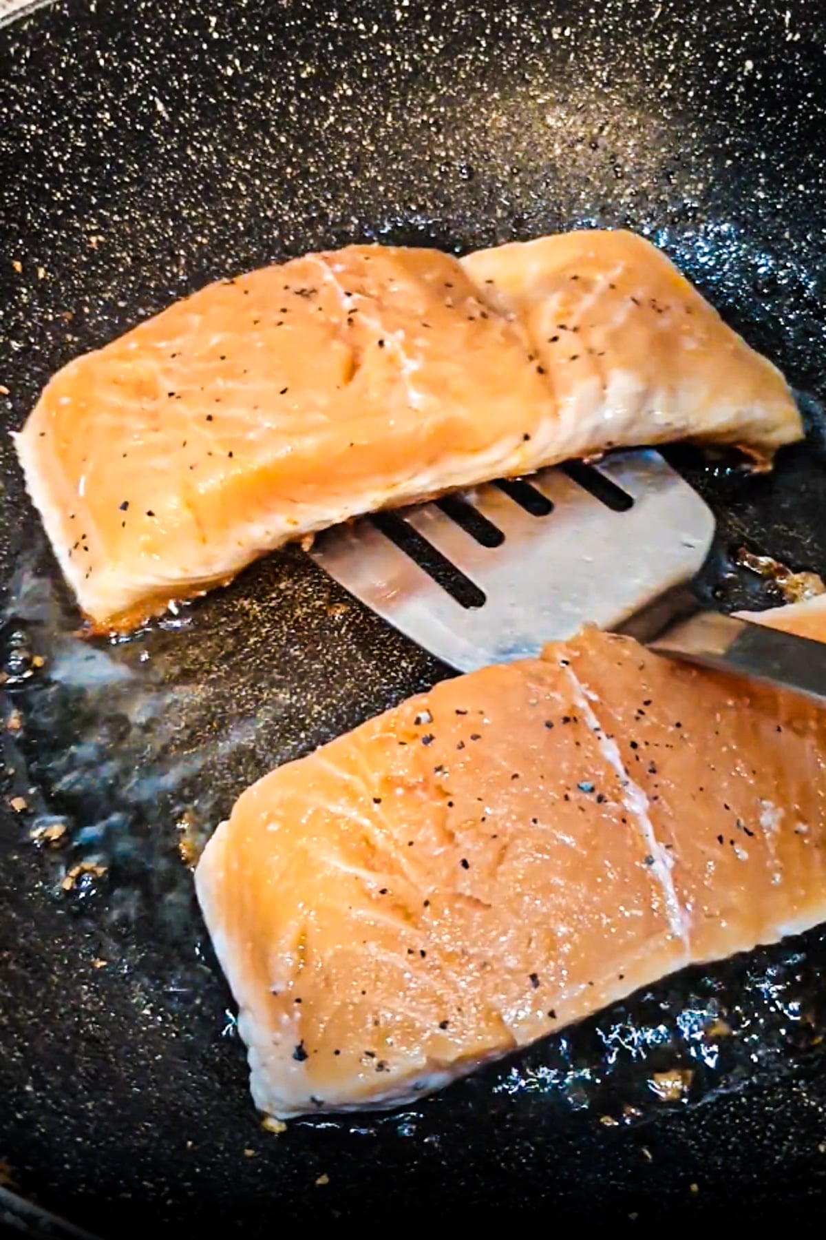 Two raw salmon fillets seasoned with pepper are being cooked in a frying pan, with a metal spatula lifting one fillet.
