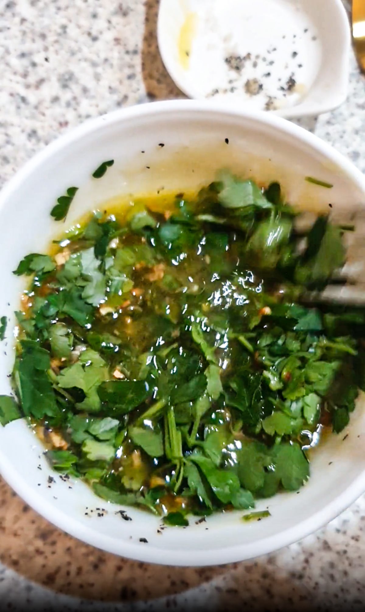 A bowl containing chopped herbs, oil, and seasonings being mixed with a fork on a speckled countertop.