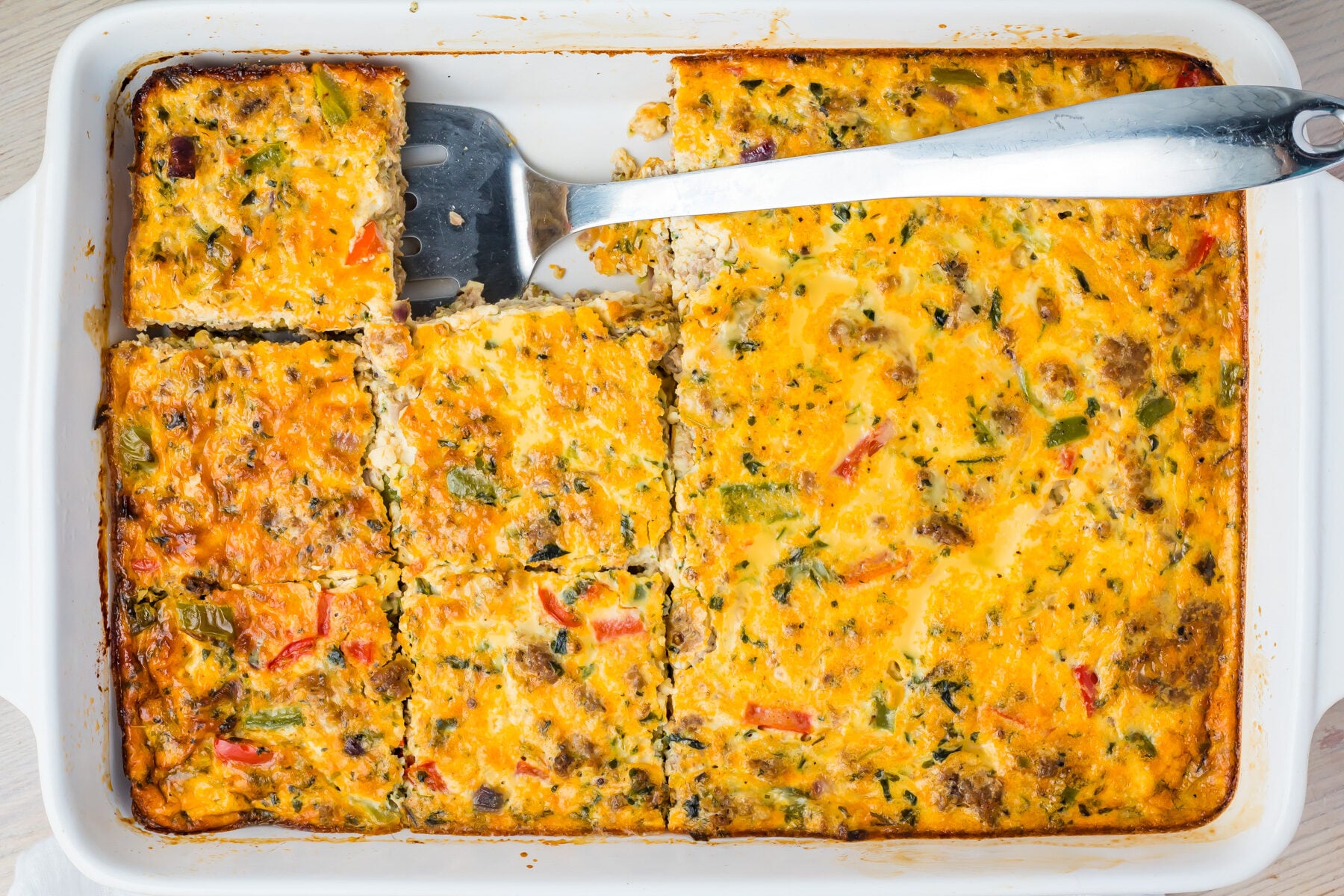 A baked sausage, egg, and cheese casserole with vegetables and sausage, sliced into squares in a white baking dish. A metal spatula rests under one of the slices.