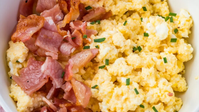 A white bowl filled with scrambled eggs garnished with chopped chives and cooked bacon pieces, set on a pink textured cloth—perfect for those seeking healthy breakfast recipes.