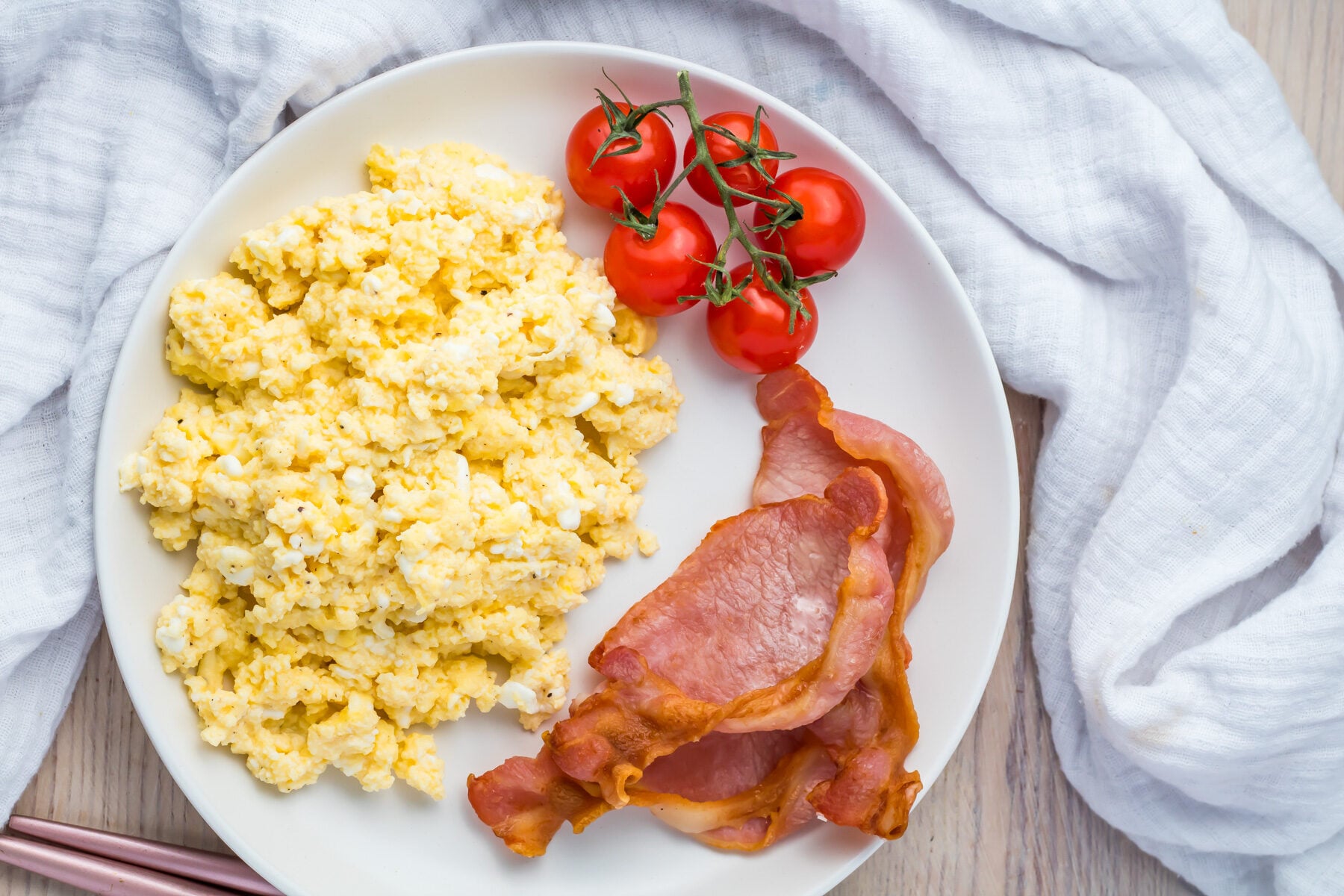 A plate with scrambled eggs, crispy bacon, and a cluster of cherry tomatoes. A white cloth napkin is partially visible in the background.