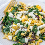 A plate of scrambled eggs with spinach, red onions, and crumbled feta cheese, served with a gold fork on a white plate.