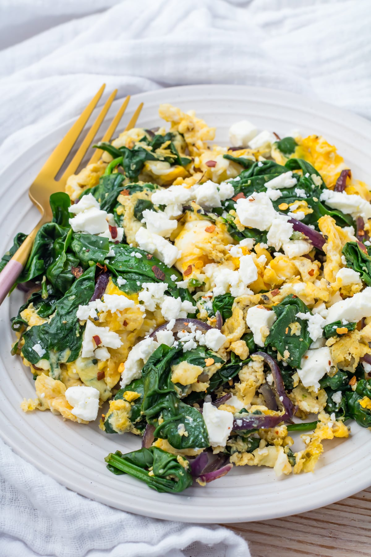 A plate of scrambled eggs with spinach, red onions, and crumbled feta cheese, served with a gold fork on a white plate.