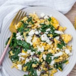A plate of scrambled eggs with spinach, red onions, and topped with crumbled feta cheese, with a fork placed on the side.